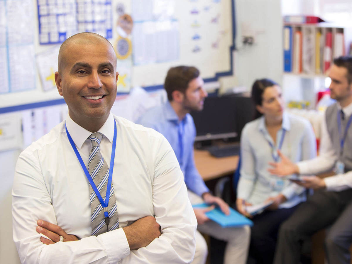 teacher smiling whilst a meeting is being conducted behind him